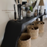 Salon bohème chic avec console Wonosobo noire en bois décorée d’objets artisanaux, panier en fibres naturelles et miroir rond.